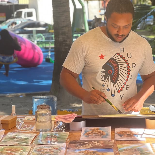 Miguel desenhando em uma feira, diante de uma mesa com suas obras